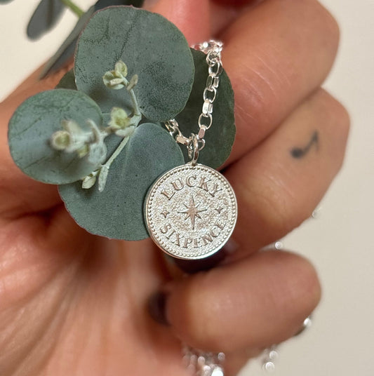 A hand holding a sterling silver lucky sixpence charm necklace entwined with eucalyptus 