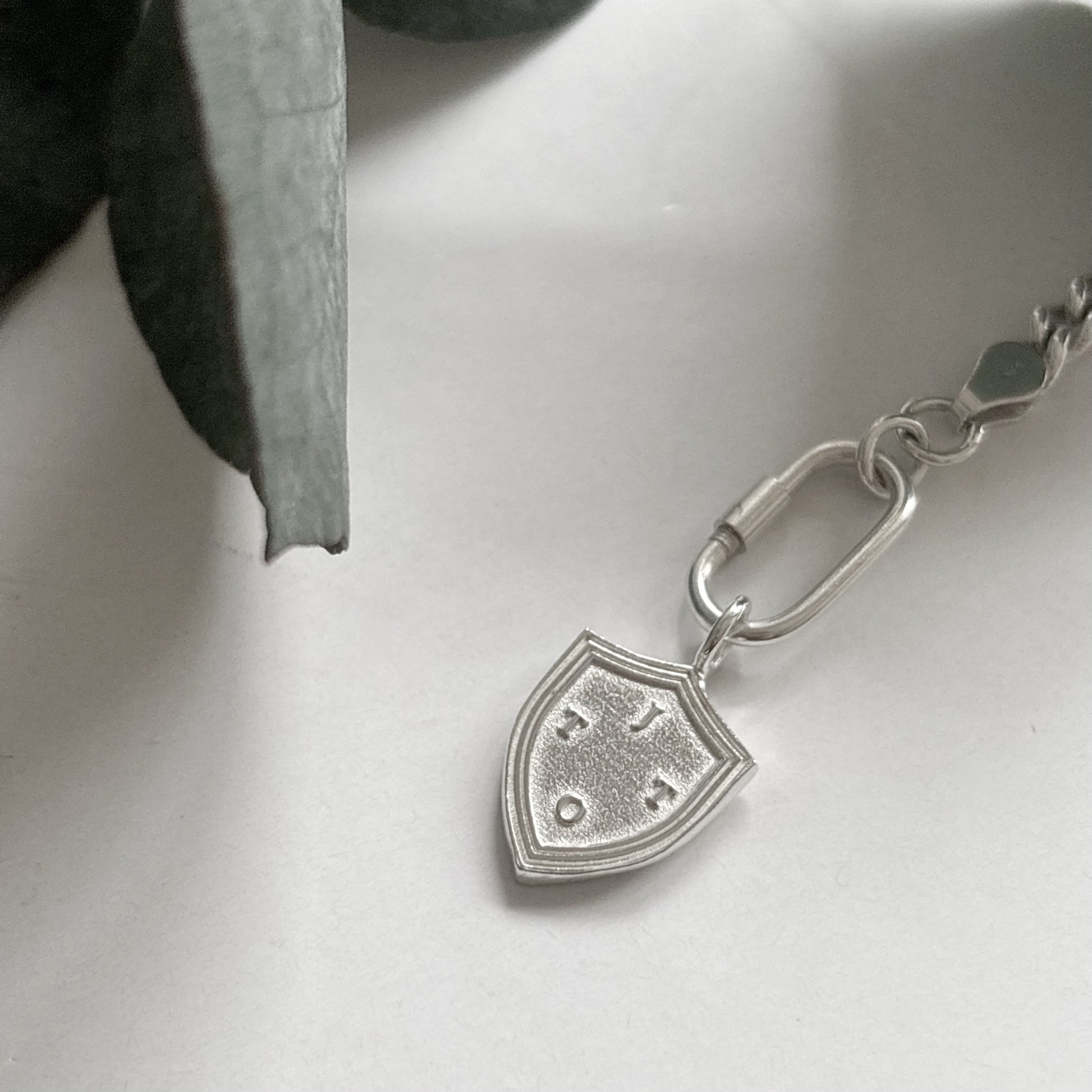 Silver Shield Charm with initials, layer on a white surface with a green eucalyptus leaf