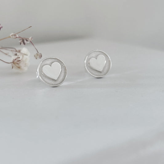 Sterling silver heart stud earrings with raised three-dimensional heart detail, photographed on a soft neutral background.
