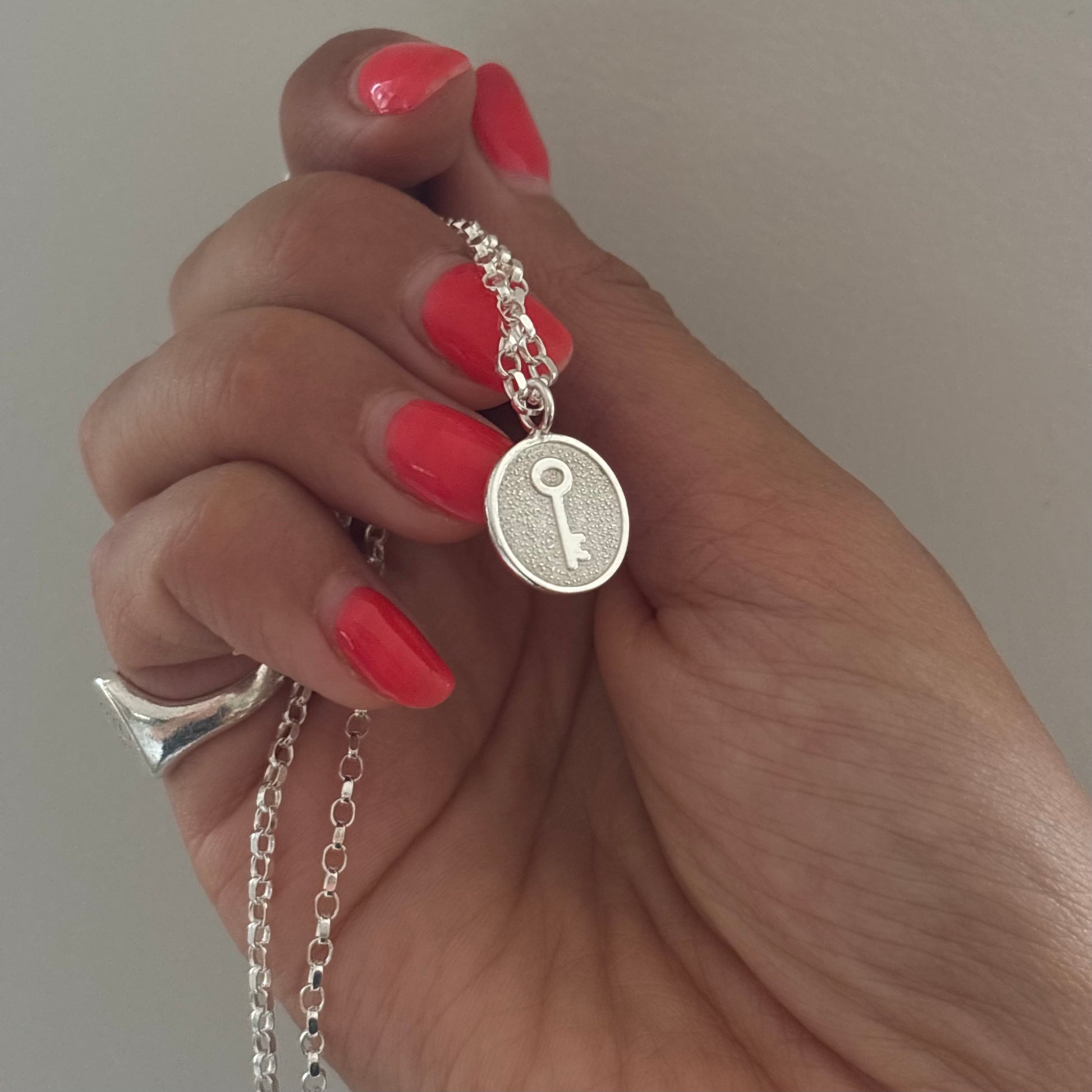 A close-up of a hand with bright coral-pink manicured nails holding a silver key pendant necklace. The pendant is round shaped with a textured background and a key design in the center, suspended from a silver chain.
