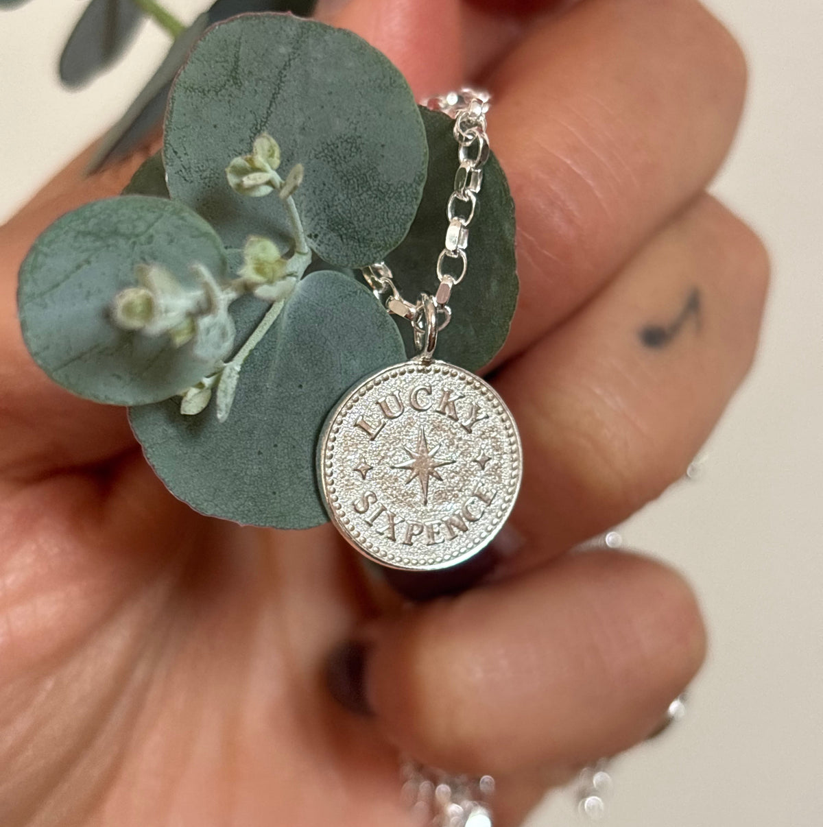 A silver coin pendant necklace engraved with 'LUCKY STUPENY' and a compass rose, held in a hand with eucalyptus leaves against a light beige background.