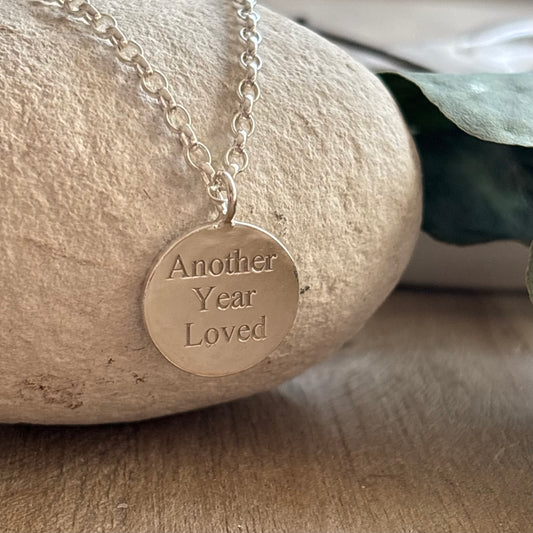 Sterling silver disc necklace engraved with the words “Another Year Loved” on a fine silver chain, styled on a stone background.