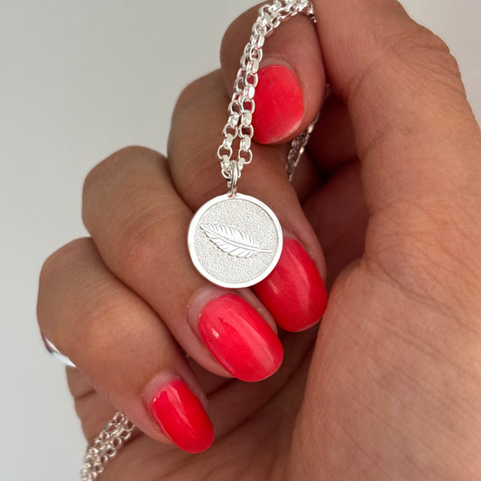 A close-up of a hand with bright red nail polish holding a silver chain necklace with a round feather pendant.