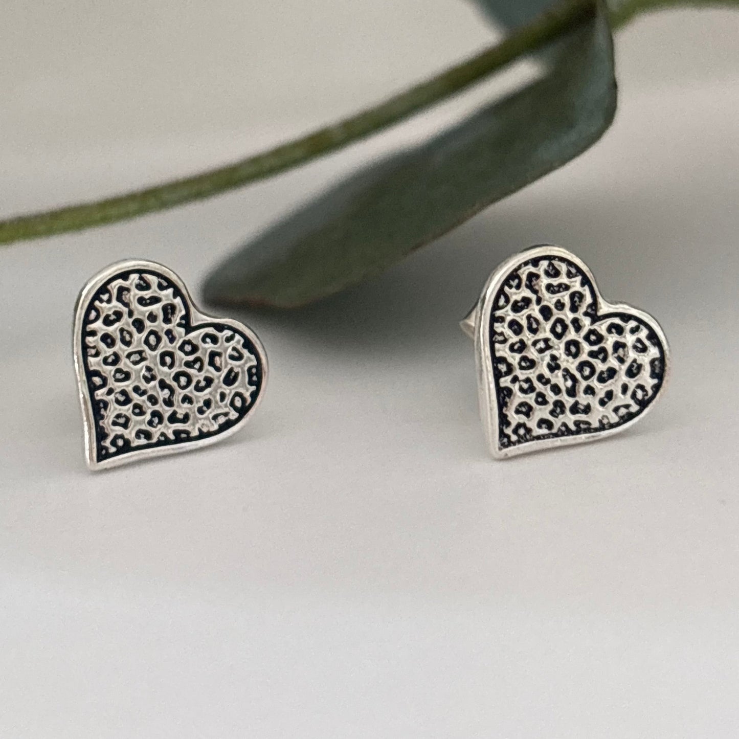 Sterling silver leopard print heart stud earrings on a white background with eucalyptus leaves
