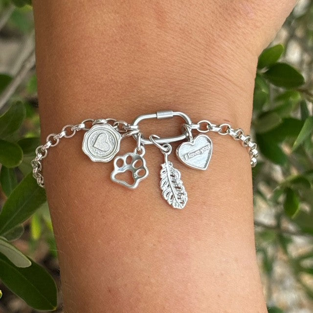 Sterling silver charm bracelet featuring four charms: a wax seal heart charm, a paw print charm, an engraved “I love you” heart charm, and a detailed feather charm, on a link chain with an oval clasp – Theodore & Olive Jewellery.
