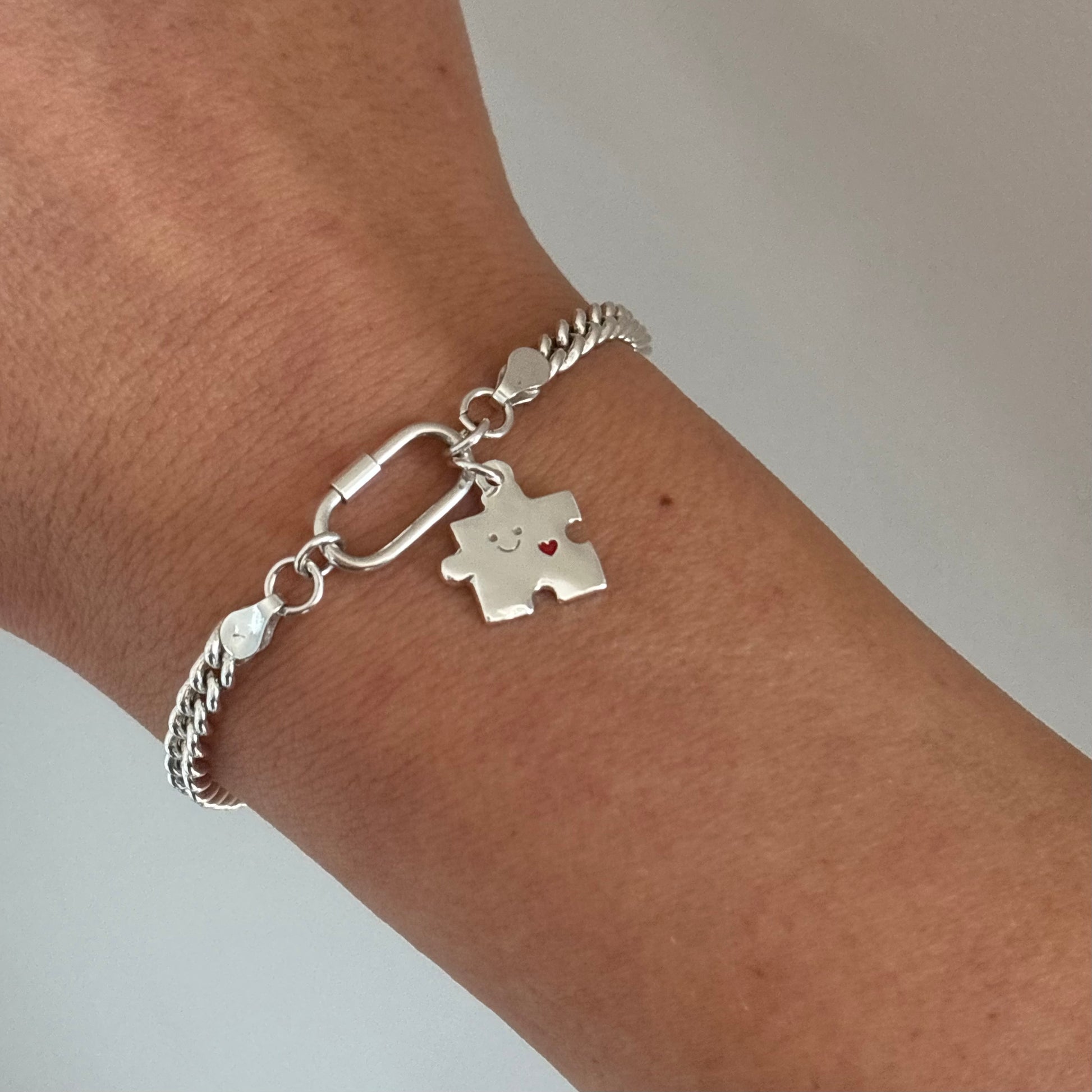 
Sterling silver bracelet with Theo’s Puzzle Charm, a small smiling puzzle piece with a red heart, worn on the wrist.