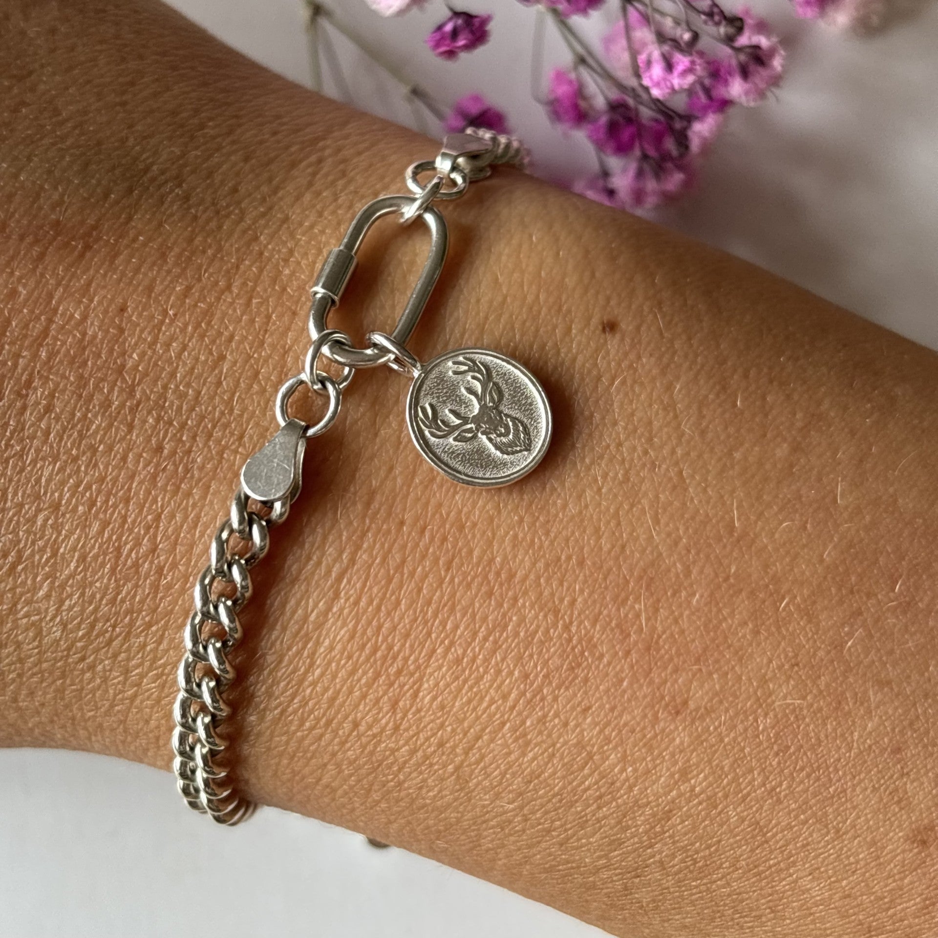 Silver bracelet with a oval stag charm on a wrist, with a blurred background of pink flowers.