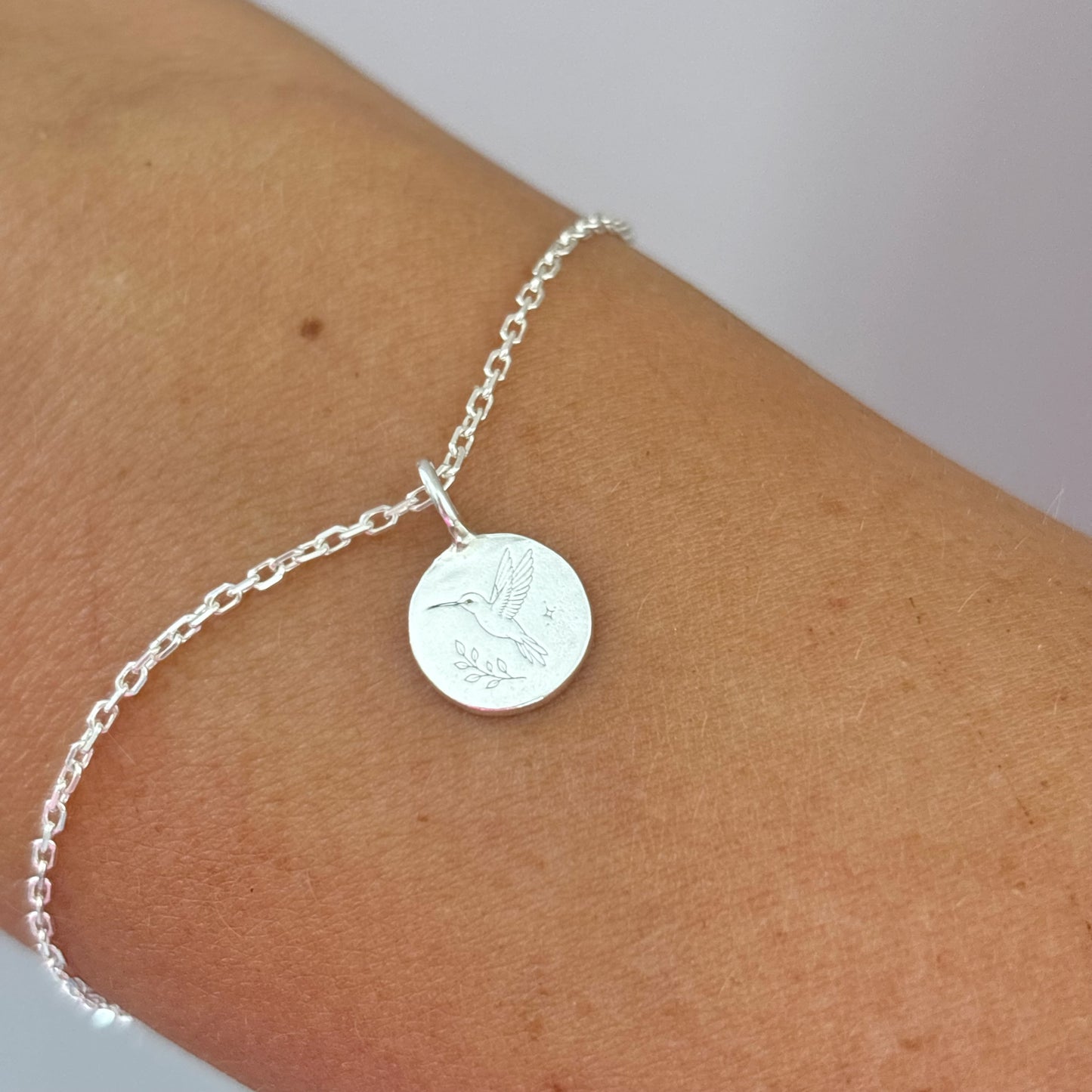 Silver bracelet with a round pendant with an engraved hummingbird on a wrist against a neutral background