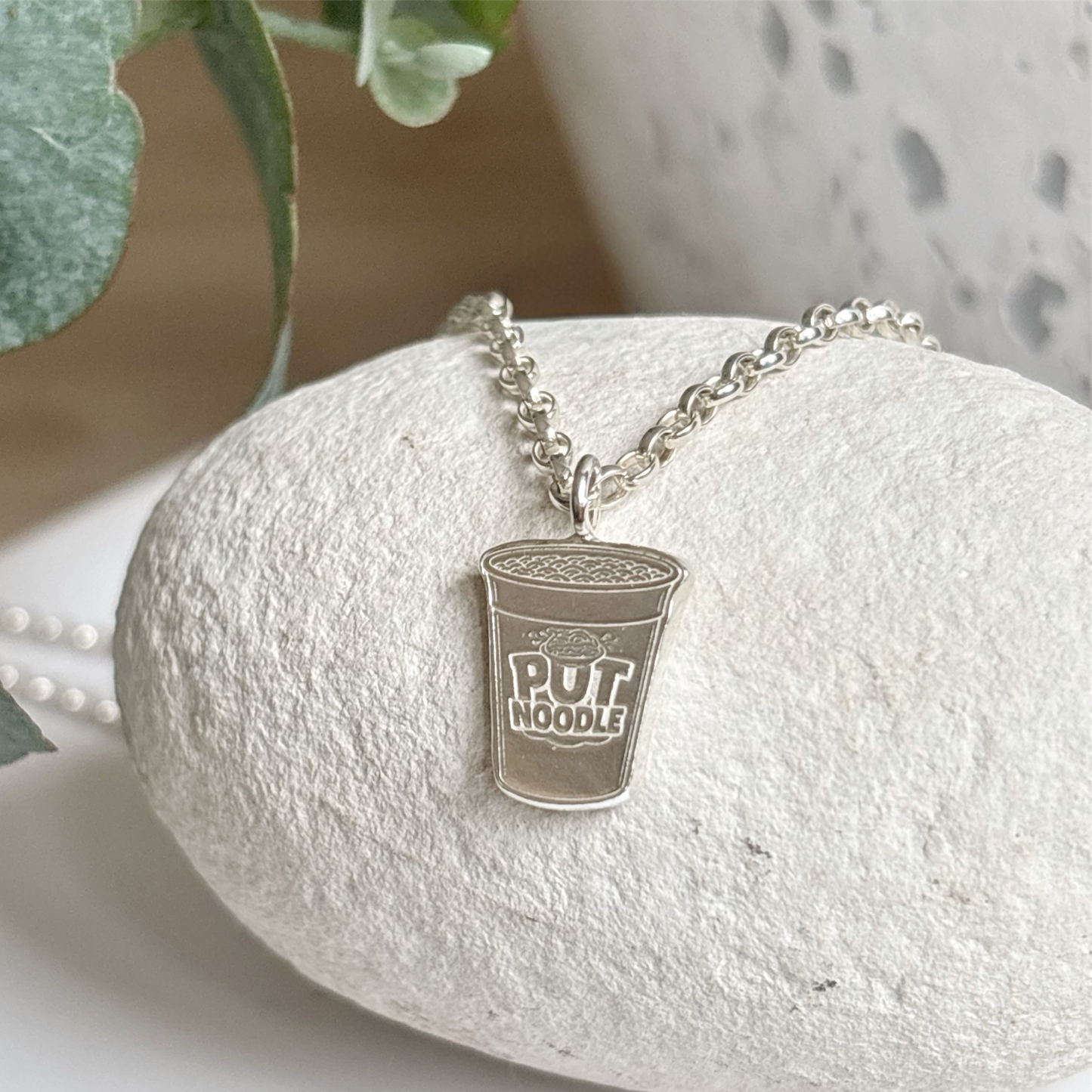 Silver pendant shaped pot noodle on a chain, placed on a white stone with green leaves in the background.