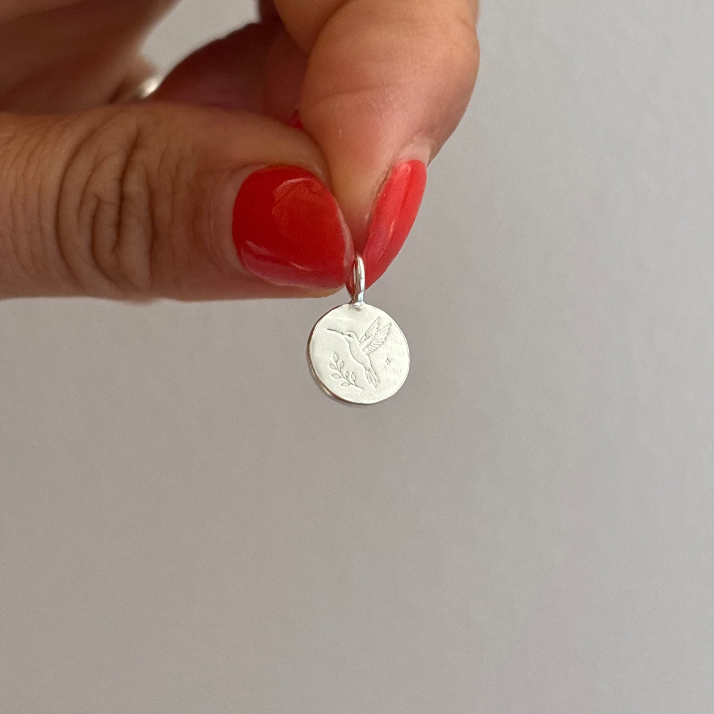 Silver pendant engraved with a hummingbird held by a hand with red nail polish on a plain background