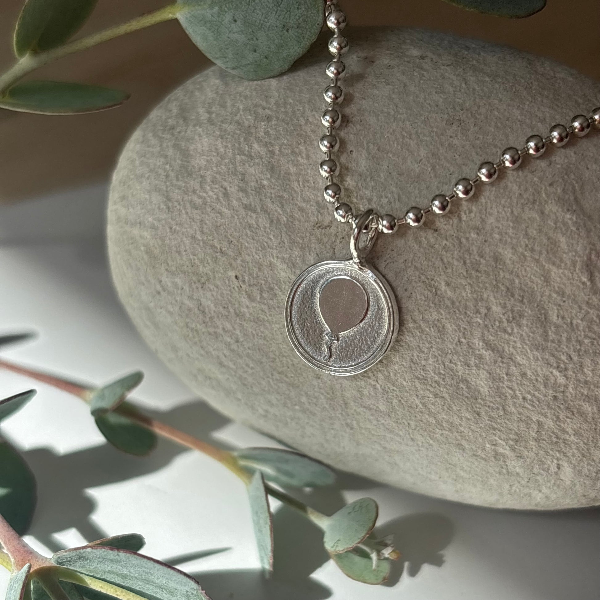 Handmade sterling silver balloon pendant necklace resting on a stone with eucalyptus leaves, meaningful nostalgic jewellery.