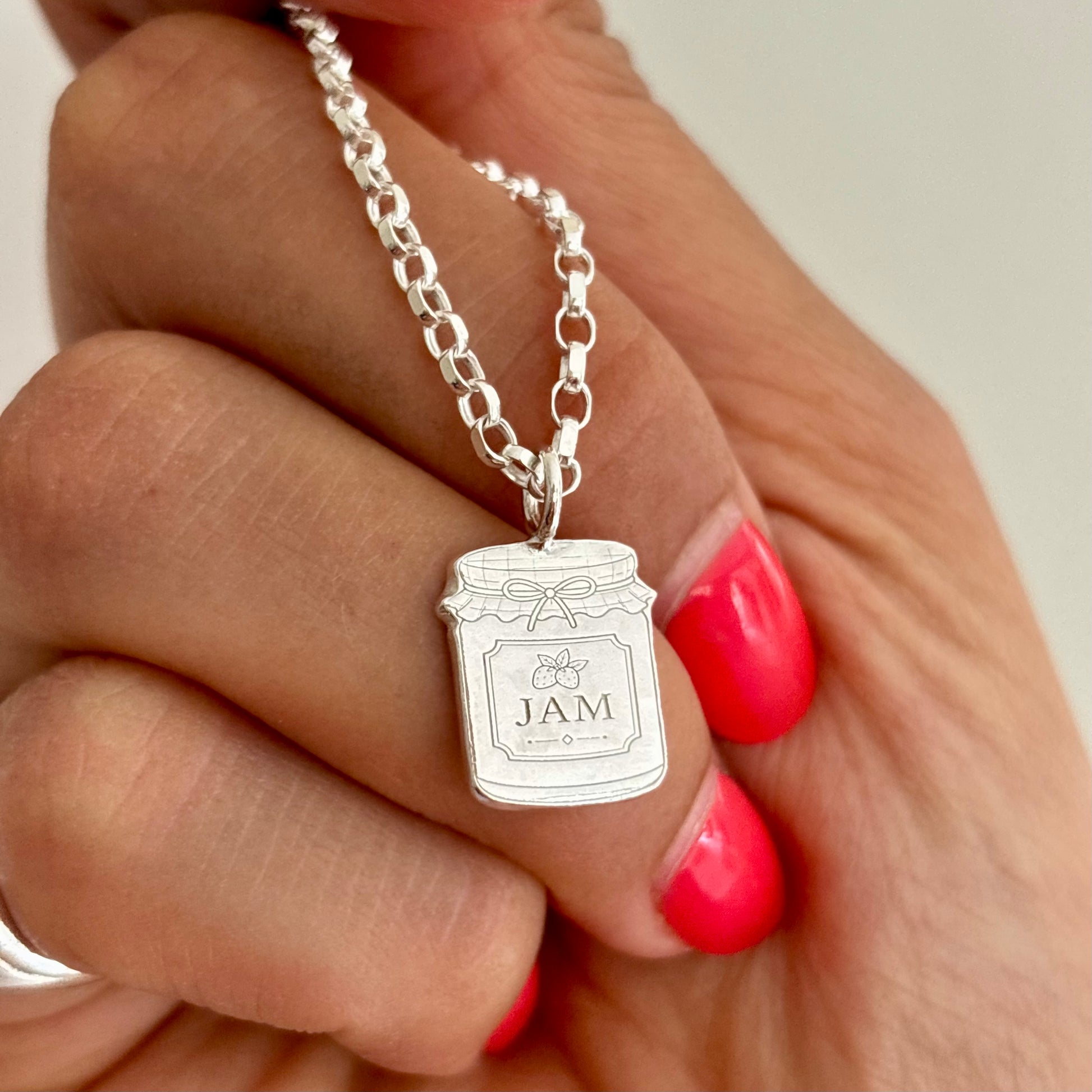 A close-up of a hand with bright pink nail polish holding a silver chain necklace with a jar-shaped pendant engraved with 'JAM' and a honeybee.