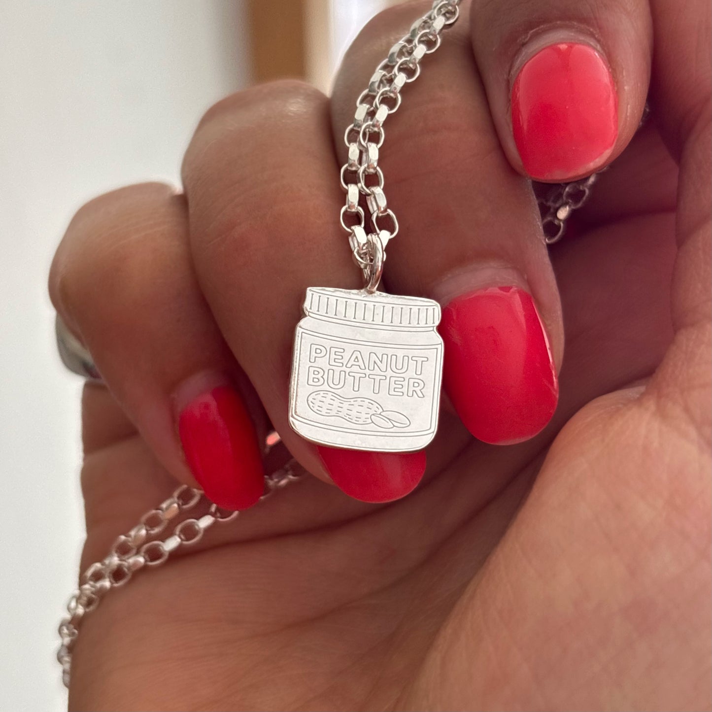 A close-up of a hand with bright red nail polish holding a silver necklace with a peanut butter jar pendant that reads 'PEANUT BUTTER'.