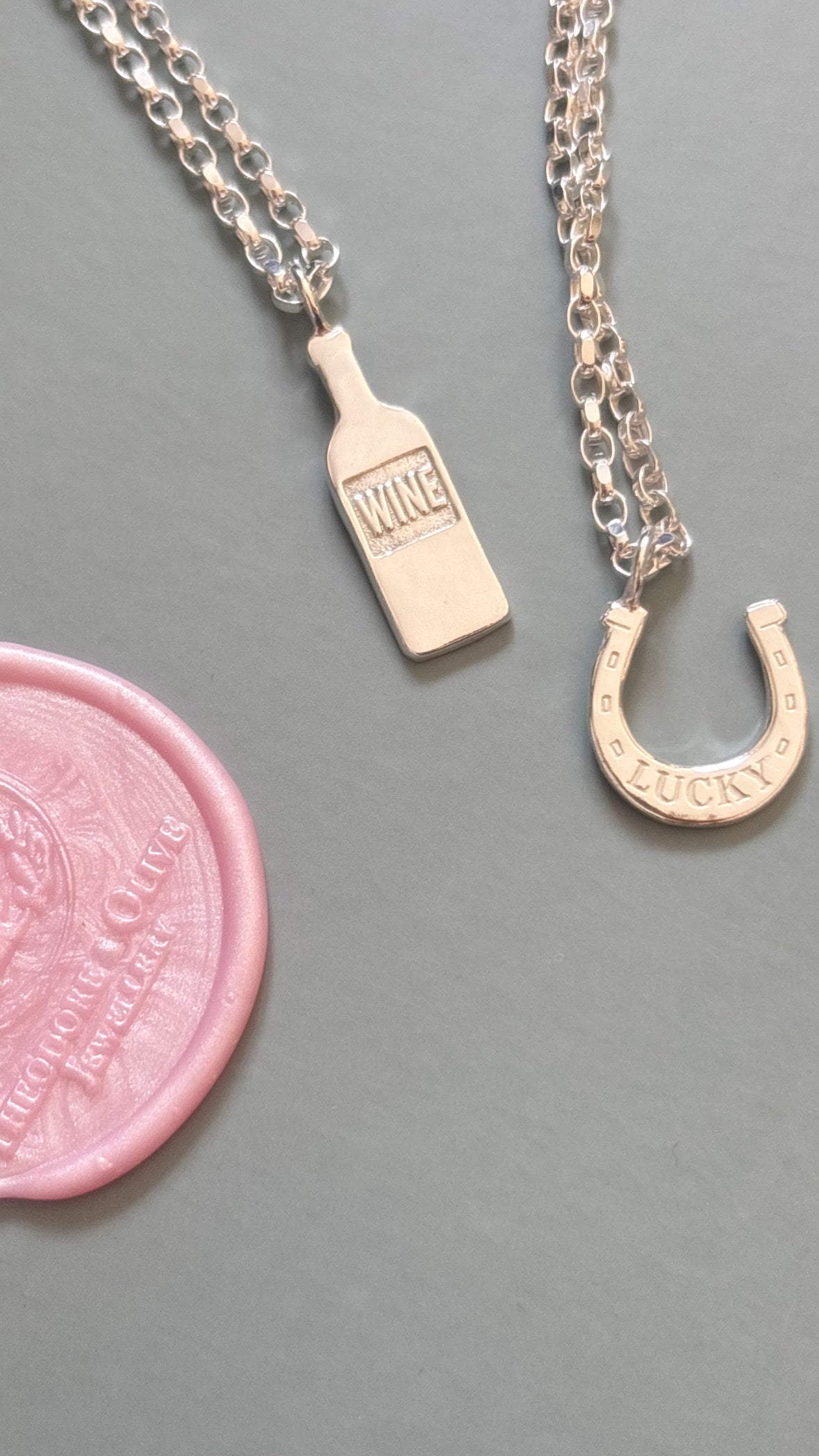 Two silver-tone chain necklaces displayed on a gray surface, one with a small bottle-shaped pendant engraved with 'WINE' and the other with a horseshoe-shaped pendant engraved with 'LUCKY'. A pink wax seal is shown at the lower left for scale.