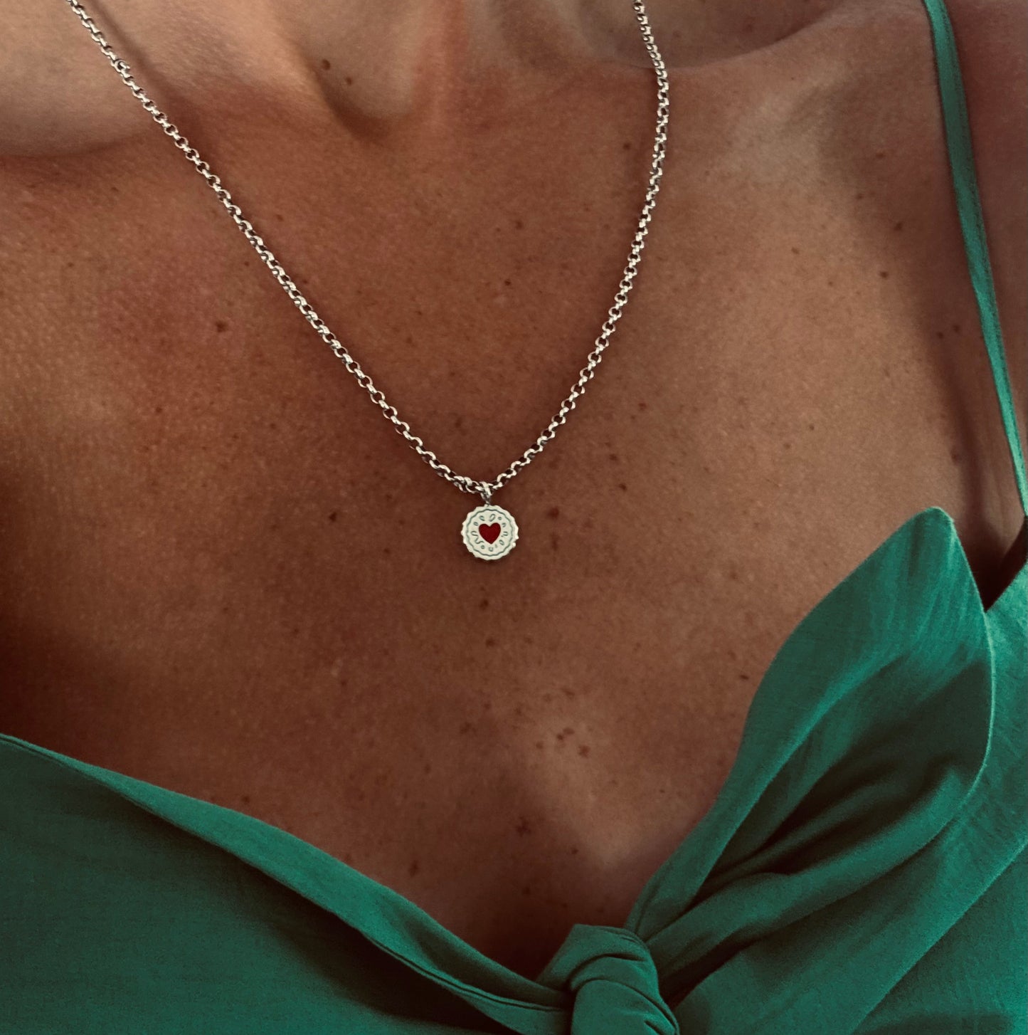 A close-up of a person wearing a delicate silver chain necklace with a Jammie Dodger pendant featuring a small red heart in the center, styled with a green tie-front top.