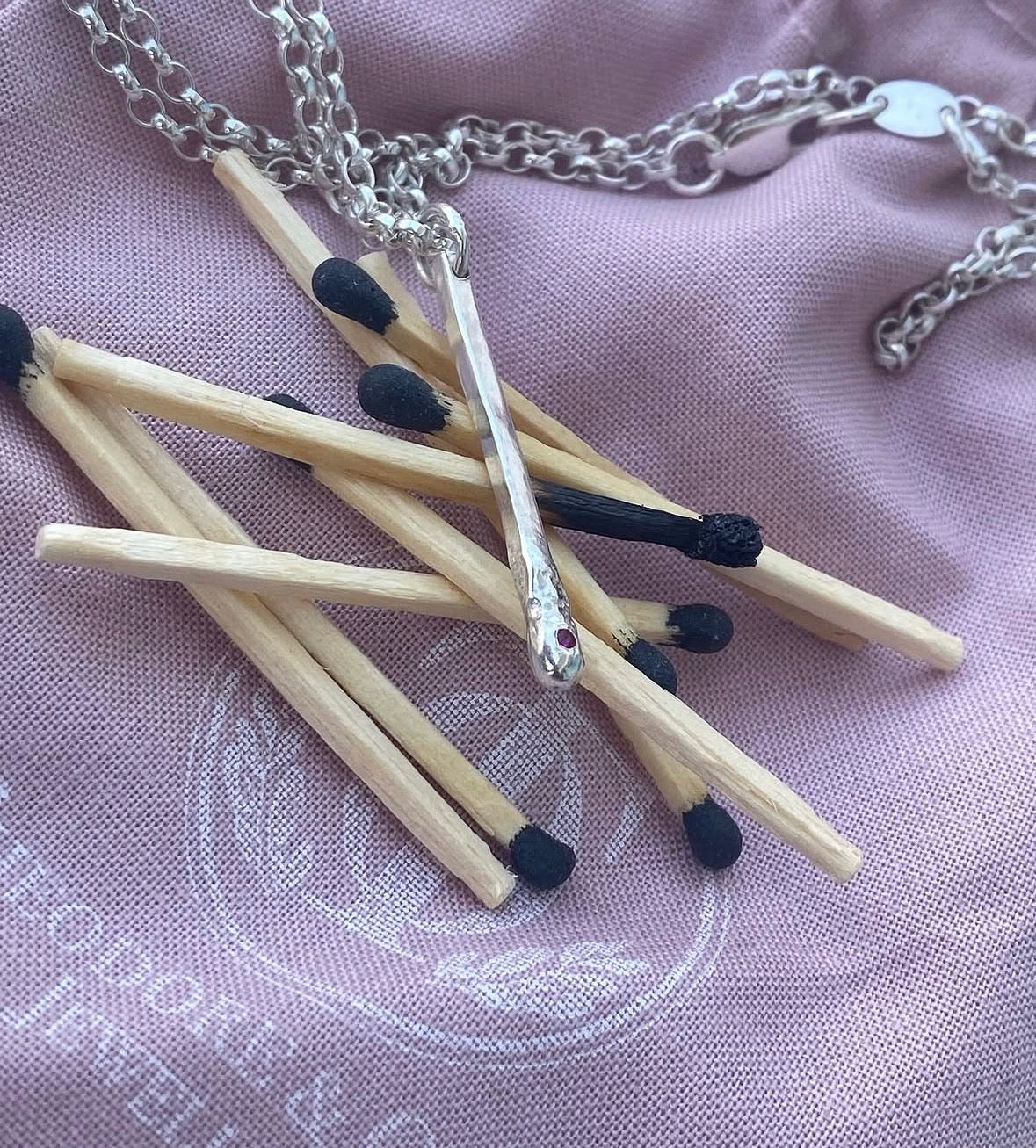 A stwrkong silver matchstick pendant necklace draped over a cluster of wooden matchsticks with black tips, displayed on a dusty lavender fabric background.