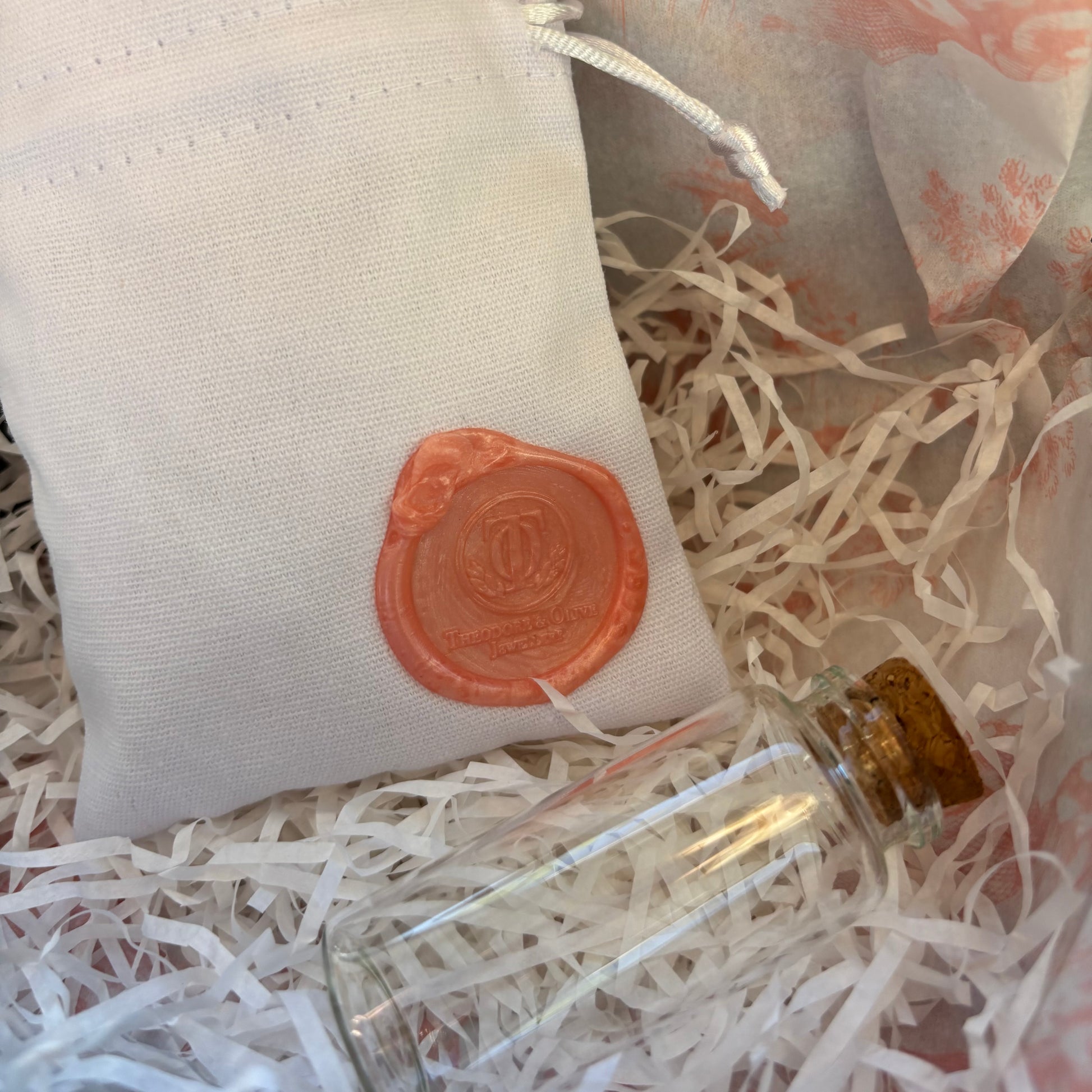 Small glass bottle with cork lid on a bed of white shredded paper, next to a beige fabric Theodore and Olive Jewellery wax seal logo pouch.