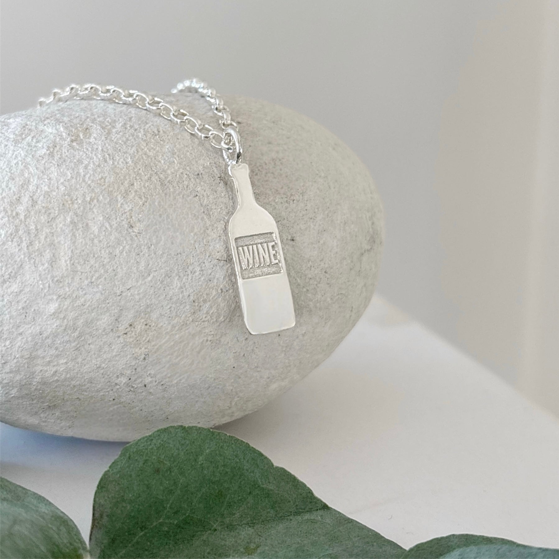 Silver necklace with a 'WINE' bottle pendant on a stone surface with green leaves.