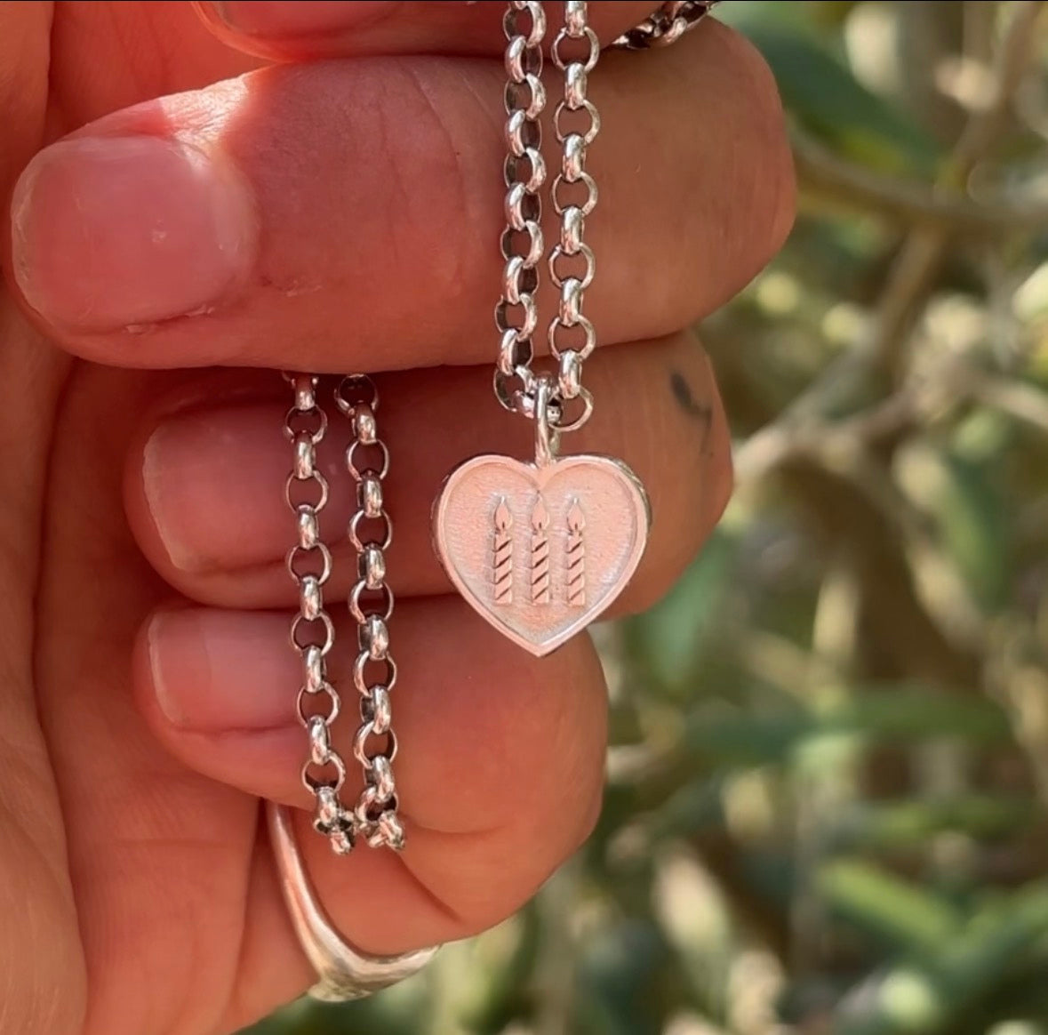 Hand holding a silver necklace with a heart-shaped pendant featuring three candles.