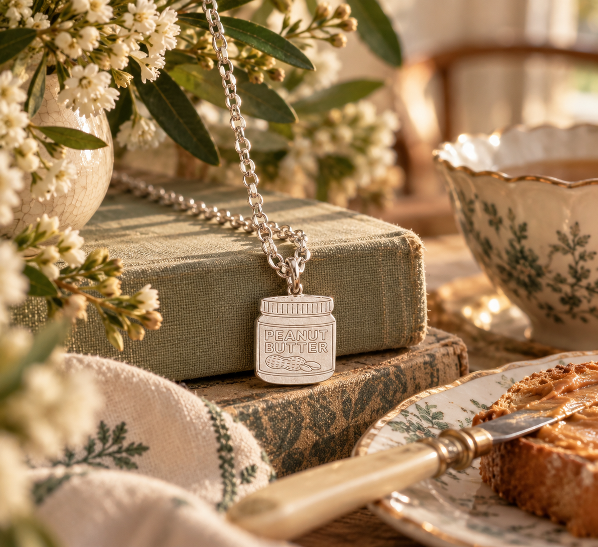 A silver-toned necklace with a jar pendant engraved 'PEANUT BUTTER FILLING' is draped over stacked vintage books in a warm, styled tabletop scene with flowers, a teacup, and toast on a plate.