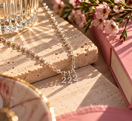 A silver chain necklace with three small dangling letters spelling '222', styled on a stone slab with pink flowers, a glass, and a pink book in warm sunlight.