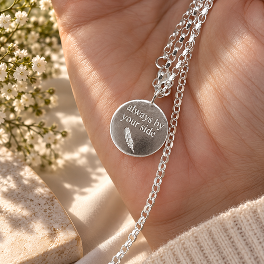 A sterling silver necklace with a round engraved disc pendant reading 'always by your side' and a feather motif, shown resting on a person's hand. The necklace features a delicate link chain and a small clear crystal accent.