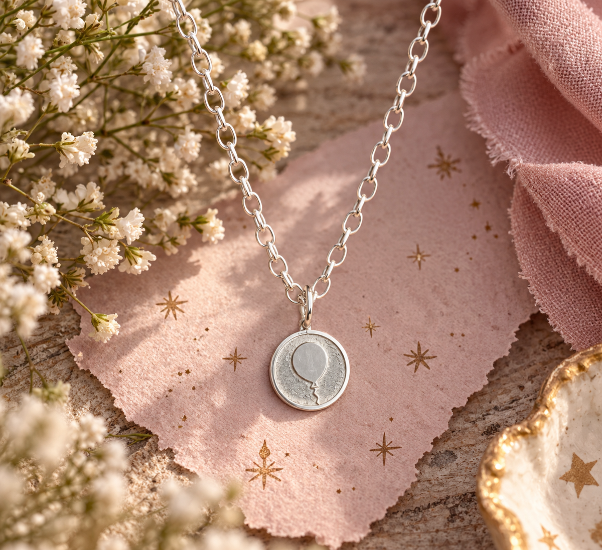 A silver chain necklace with a round pendant featuring a balloon design, styled on a pink textured card with gold star prints, surrounded by white baby's breath flowers and a soft pink fabric.