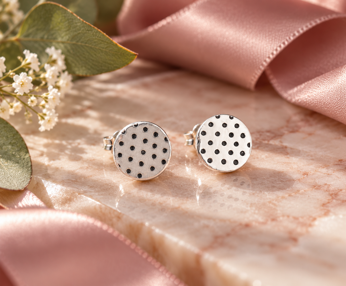 A pair of round silver-toned earrings with a white background and black polka dots, styled on a pink marble surface with a pink satin ribbon and baby's breath flowers.