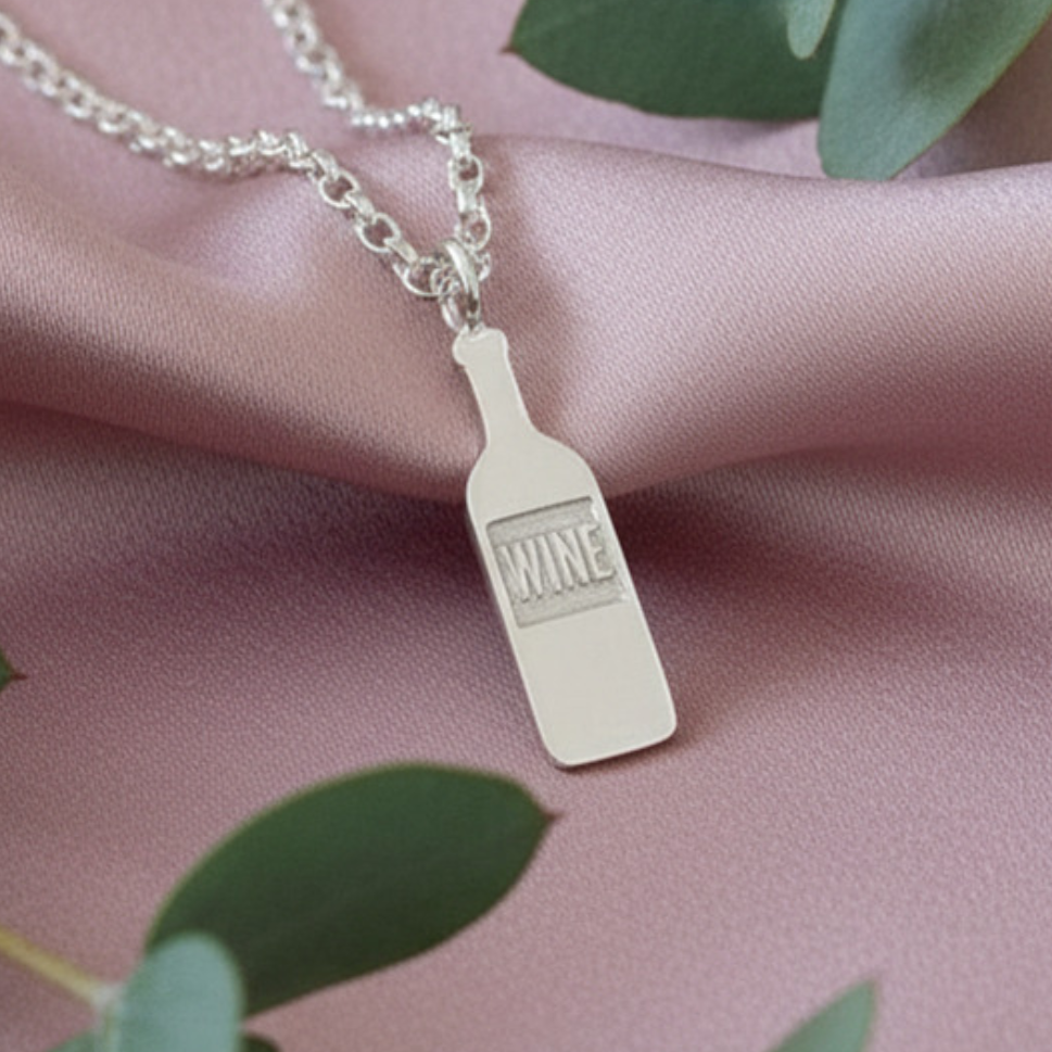 Necklace with a wine bottle pendant on a pink fabric background with green leaves.