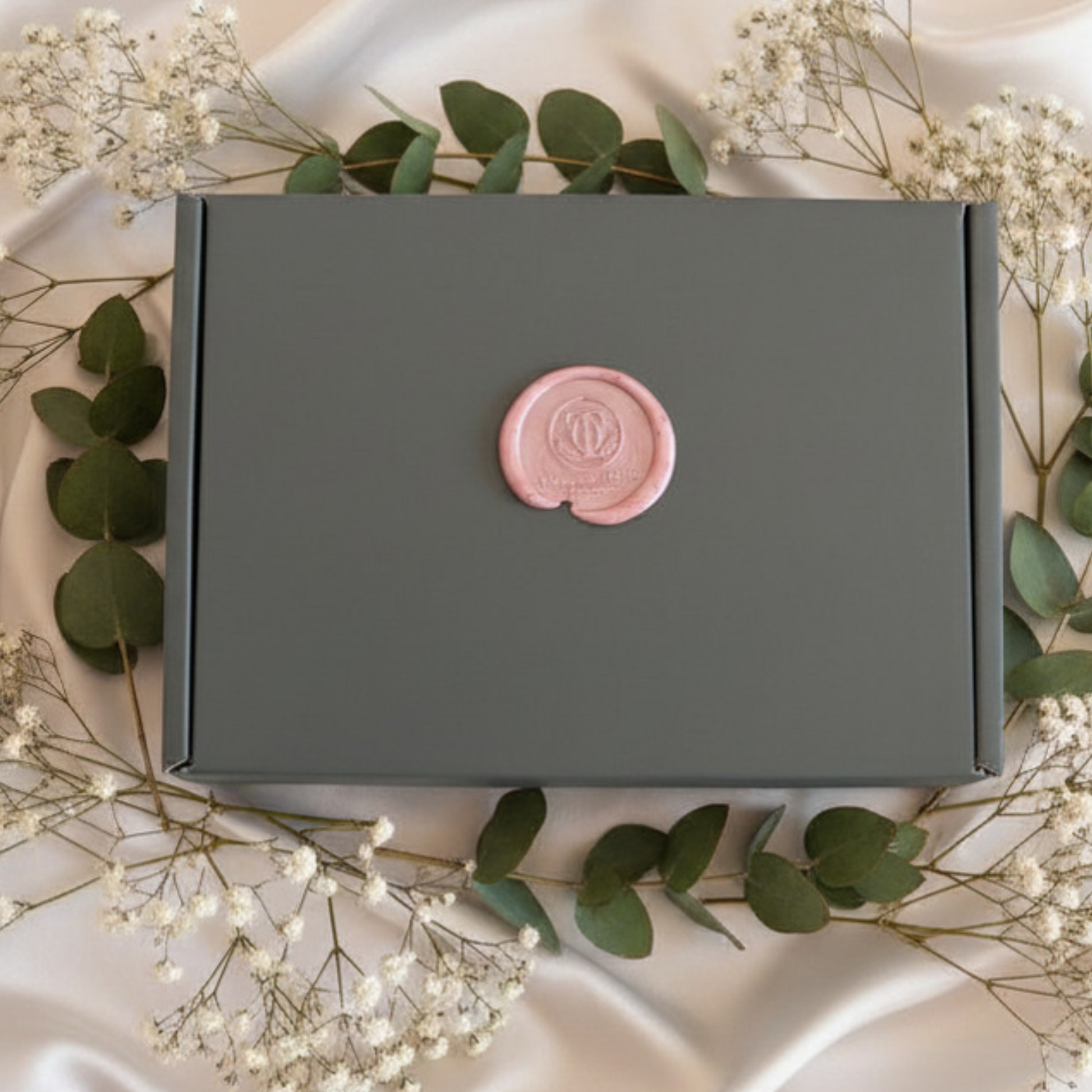 Green box with a pink wax seal surrounded by greenery on a beige fabric background