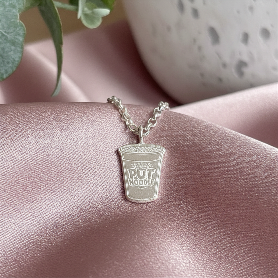 Silver pendant shaped like a cup with Pot Noodle text on a pink surface