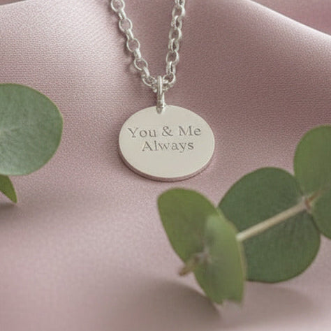Silver necklace with a round pendant on a pink fabric background with green leaves.