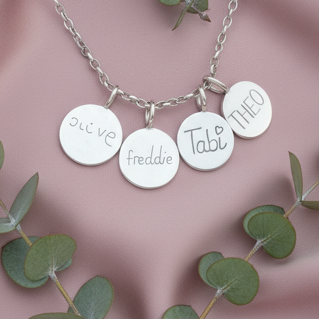 Silver necklace with engraved charms on a pink fabric background with eucalyptus leaves.