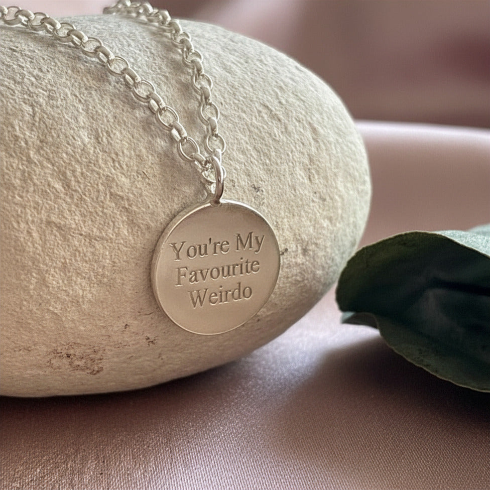 Necklace with a silver pendant reading 'You're My Favourite Weirdo' on a stone surface.
