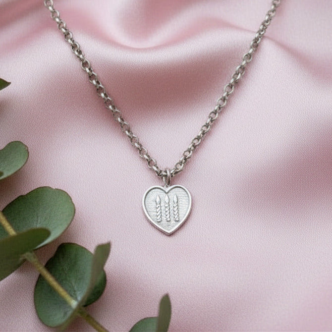 Silver necklace with a heart-shaped pendant on a pink fabric background with green leaves.
