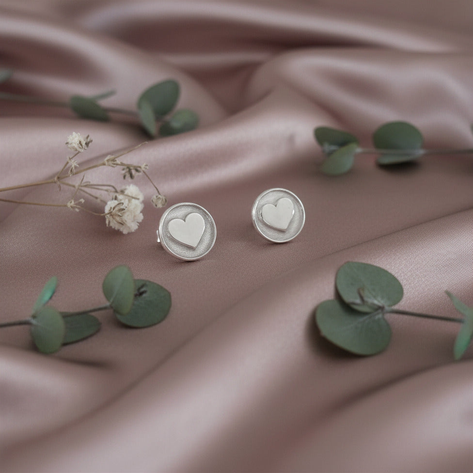 Silver heart-shaped earrings on a soft pink fabric background with green leaves and white flowers.