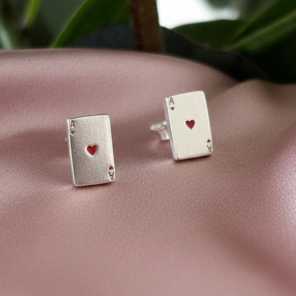 Silver playing card earrings with a red heart on a pink surface with green leaves in the background