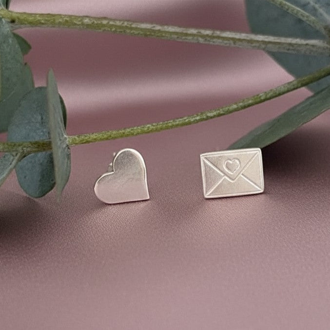 Silver heart and envelope earrings on a pink surface with green leaves.