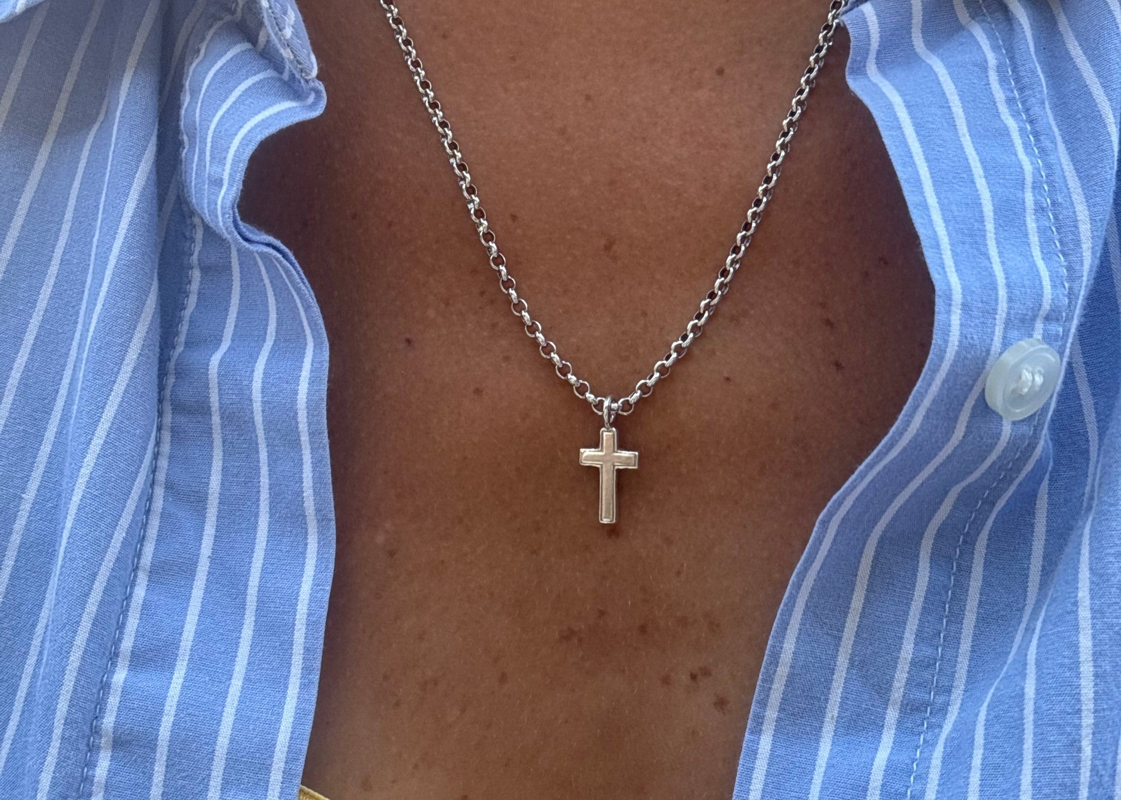 A close-up of a person wearing a silver cross pendant necklace over a blue and white striped button-down shirt.